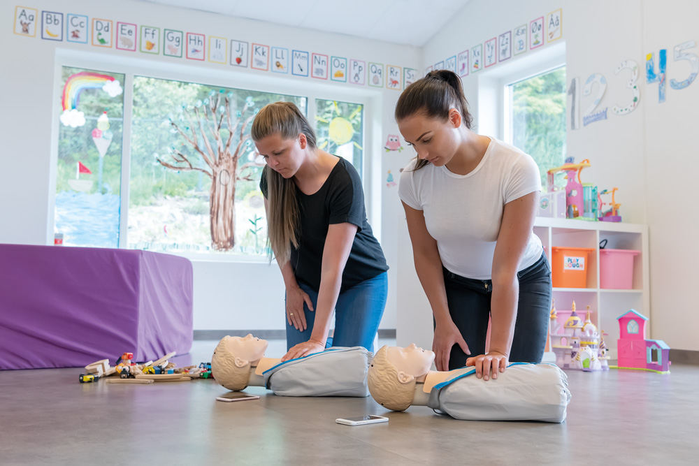 Två kvinnor övar HLR på Little Junior QCPR-dockor i en färgglad förskolemiljö.